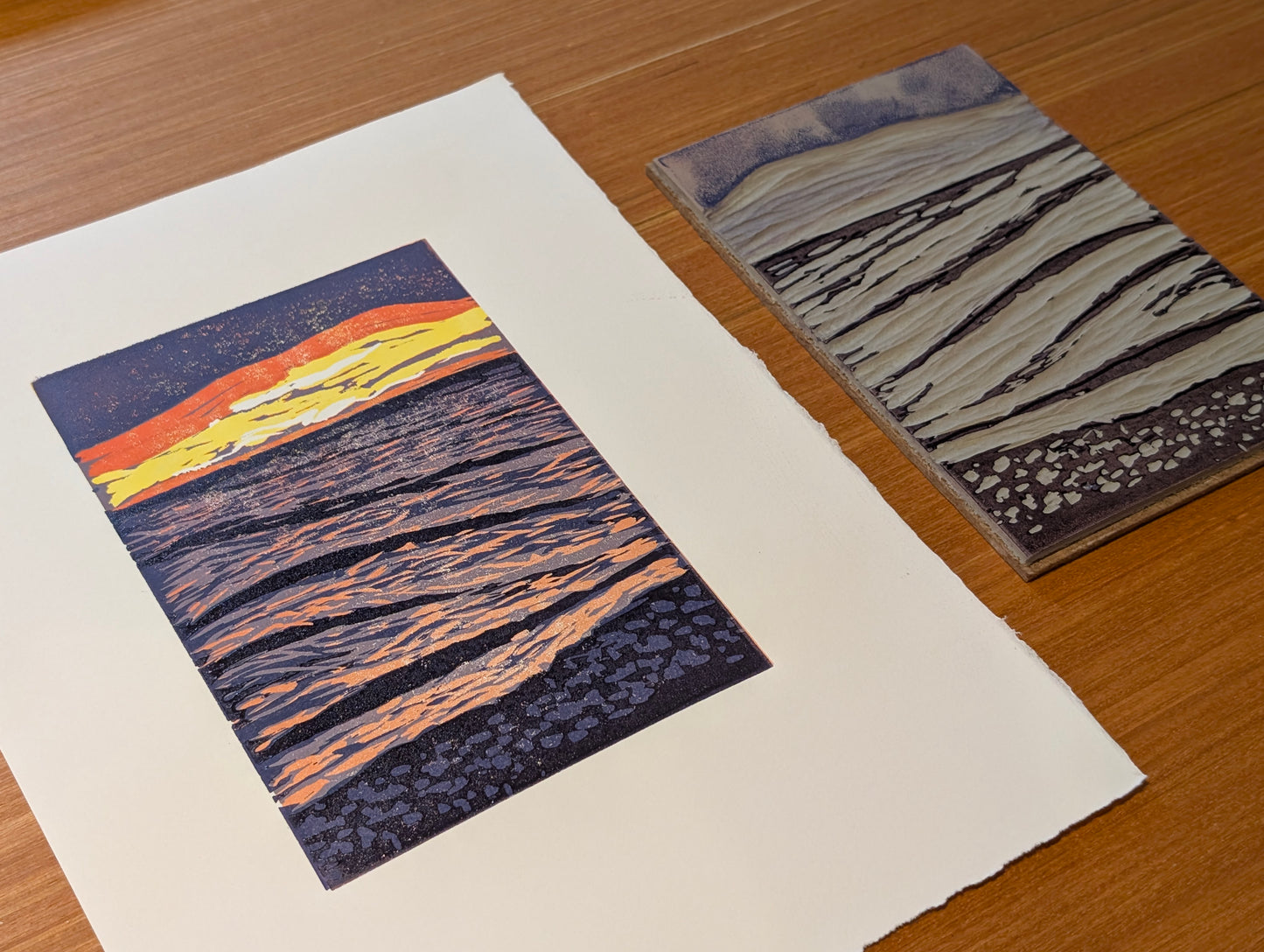 An art print showing a stylized sunset over rippled water, displayed next to the lino block on a wooden tabletop.