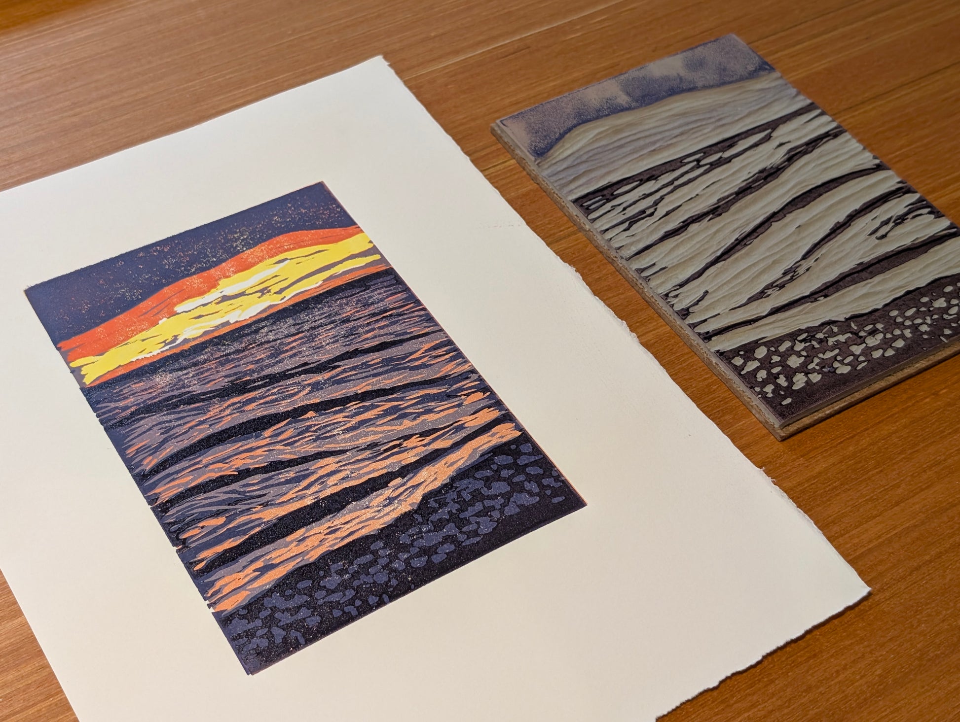 An art print showing a stylized sunset over rippled water, displayed next to the lino block on a wooden tabletop.