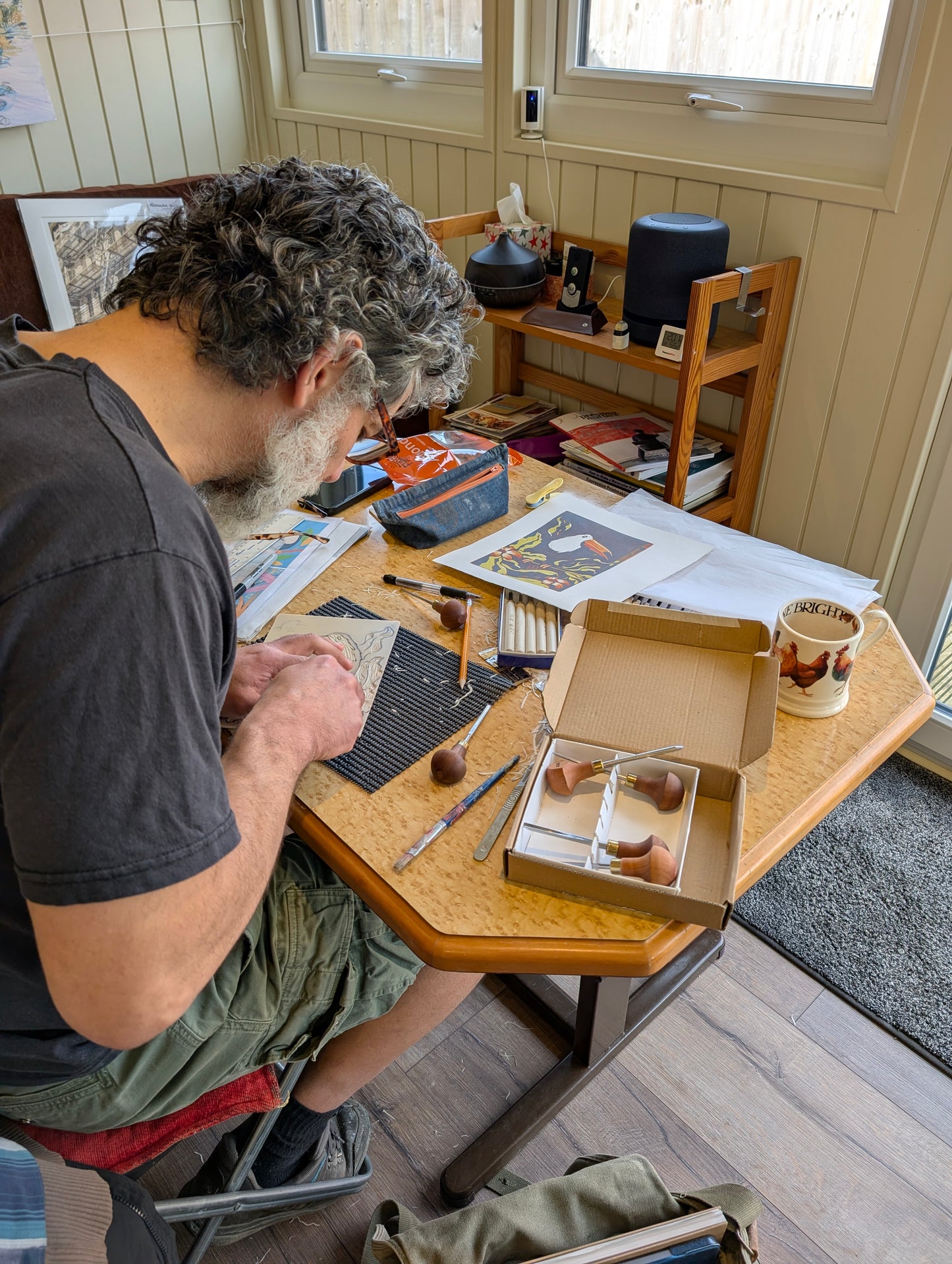 A man with curly gray hair and a beard sits at a wooden table, carving lino. The table is covered with carving tools, papers, a mug, and an open box containing several lino gouges.