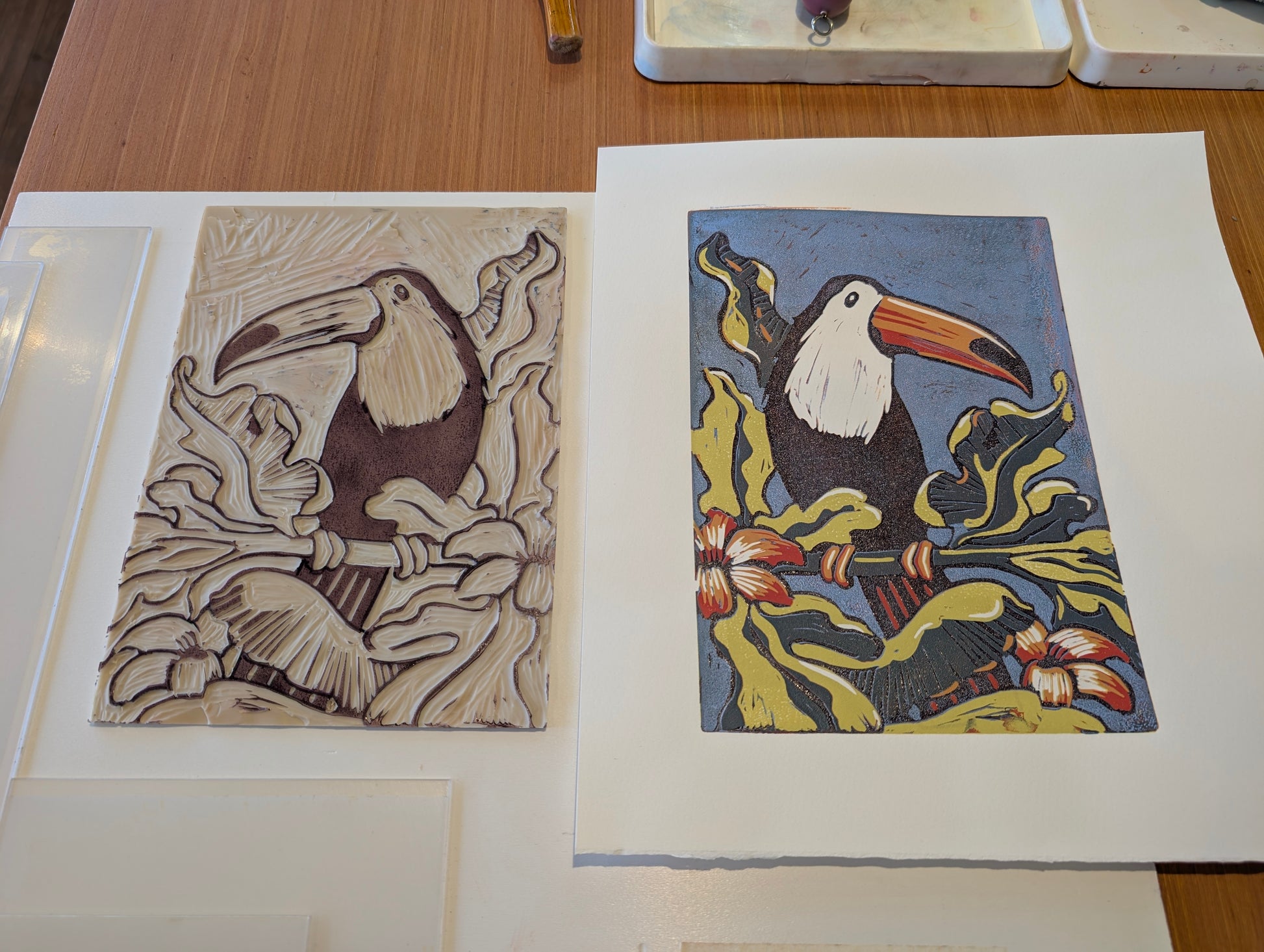 A colourful toucan lino print next to the lino block which create it on a wooden table.