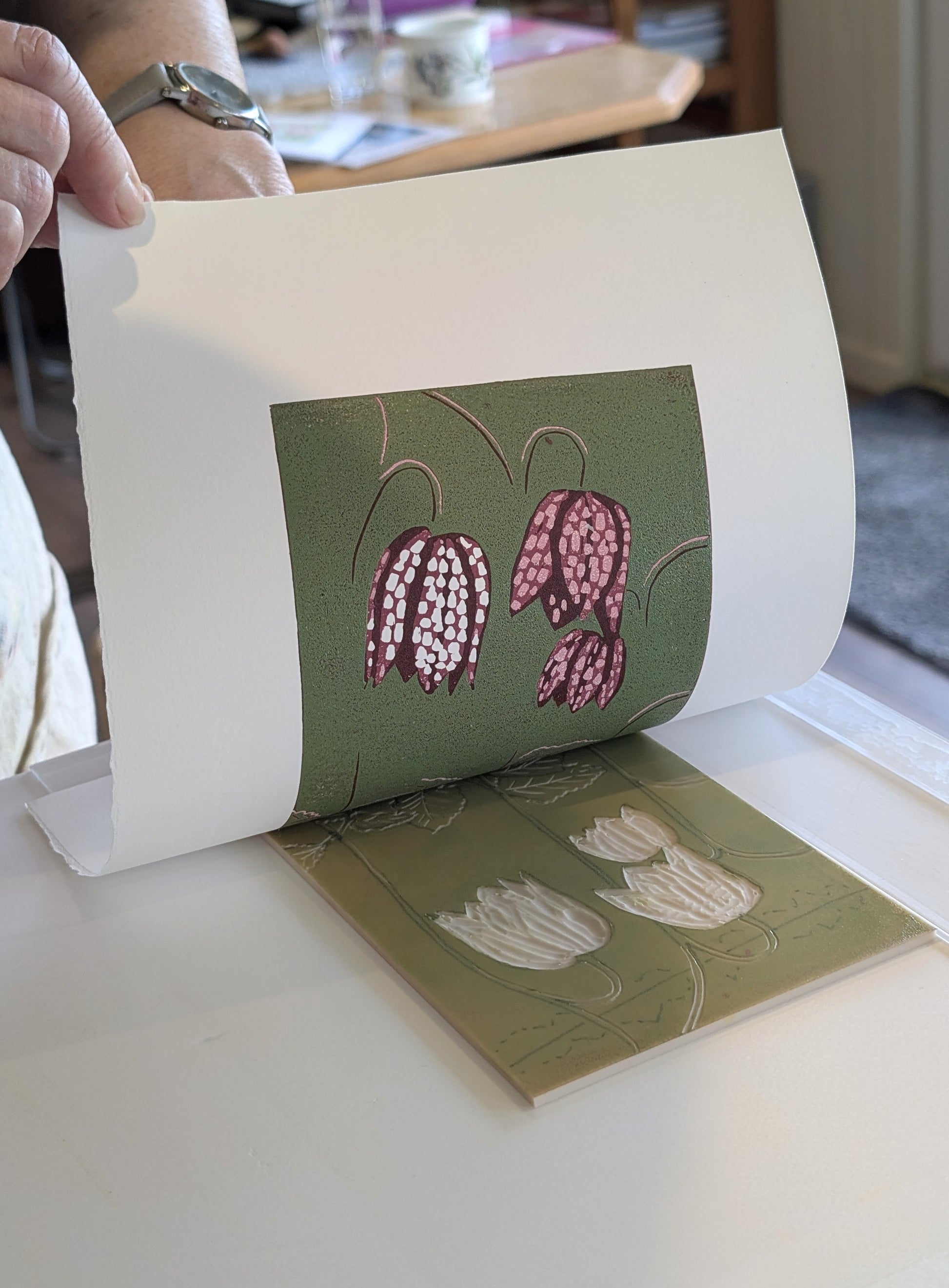 A person's hand peels a floral print from the lino.  Pink-and-white hanging flowers on an olive green background. 