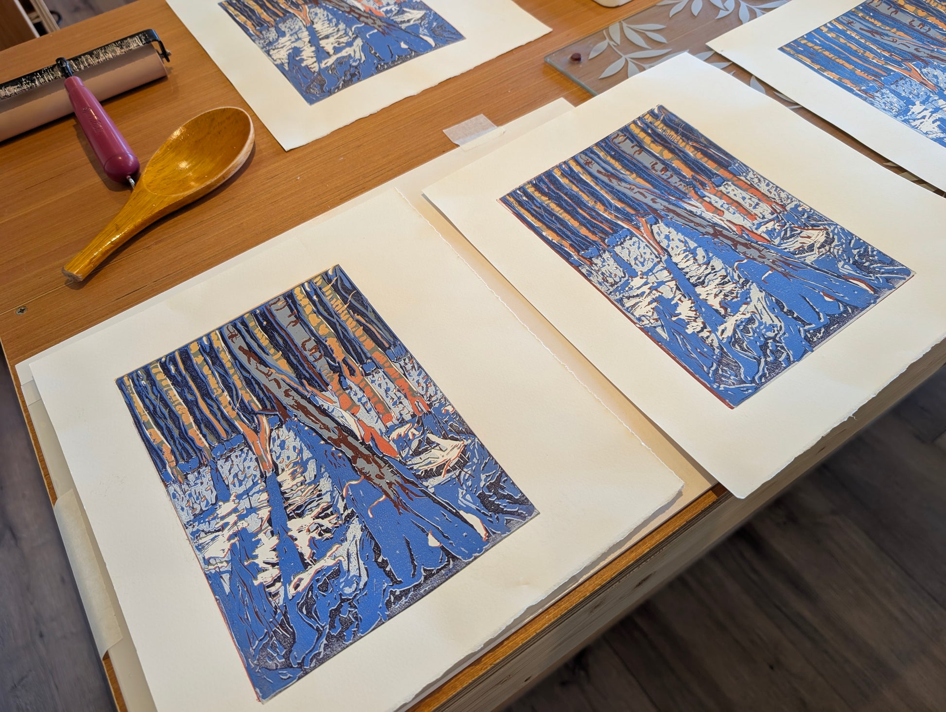 A close-up of several printed art sheets laid out on a wooden table. The sheets feature a stylized winter forest scene with blue water, white snow, and tan/orange tree trunks. Lino printing brayer and spoon in background.