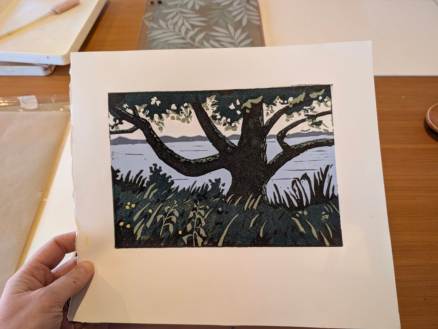 A hand holds an art print featuring a block-print style illustration of a large tree by a body of water, shown on a wooden table.