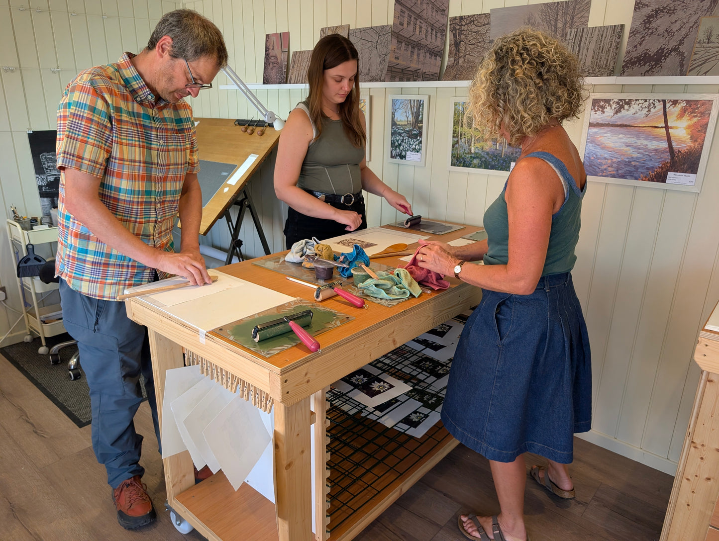 Three people gathered around a wooden printing table in a studio, working with paper, ink, and printing tools. The room has light wood paneled walls with framed artwork hanging above the workspace.