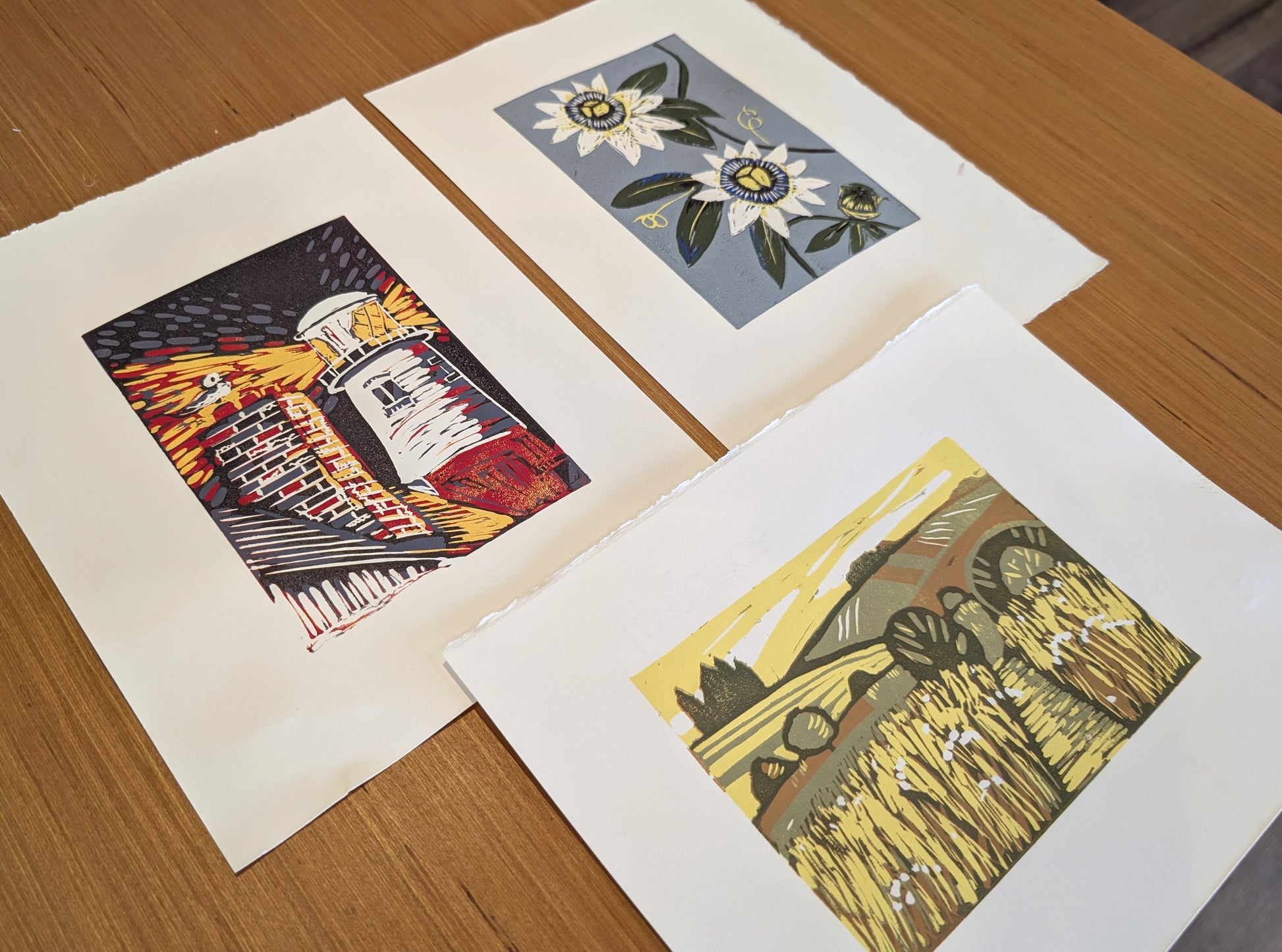 Three multi-colored lino art prints with white margins are arranged on a wooden table. The prints depict a colorful lighthouse, white flowers on a blue-gray background, and a yellow wheat field.