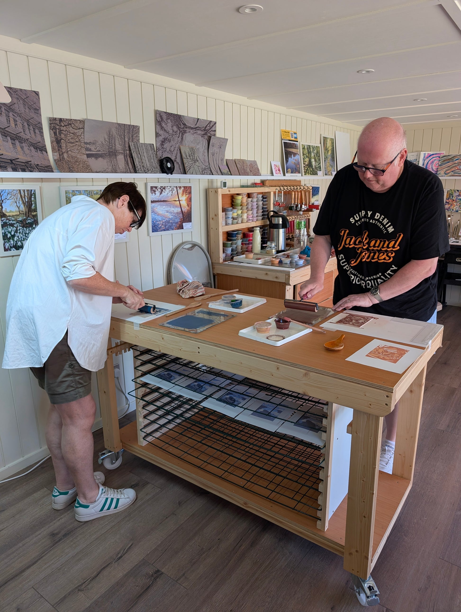 Two people working at a wooden art table inside a bright print studio, with sketches, papers, lino blocks and shelves visible in the background.